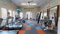 This fitness center is designed to inspire and energize its users from the moment they walk in. The space features a lively, motivational atmosphere, with bold typography murals covering the walls in teal, orange, and red hues, displaying words such as "Energize," "Train," "Focus," and "Empower." Colorful modular carpet tiles in shades of teal, orange, and gray add a dynamic visual element to the floor, complementing the vibrant wall décor.

The room is bright and airy, benefiting from multiple large arched windows that allow generous natural light to fill the space. Ceiling fans keep the environment comfortable during workouts. A wall-mounted flat-screen television is positioned for easy viewing from the cardio equipment area.

The fitness floor is well-stocked with a variety of cardio machines, including treadmills, ellipticals, and stationary bikes, all arranged in an orderly row along one side of the room. On the opposite side, a comprehensive cable and weight machine system, along with additional strength training equipment, provides options for resistance and functional training. A large mirror along one wall further enhances the sense of openness. Overall, the space offers a complete and motivating fitness environment suitable for a wide range of workout routines.