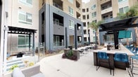This expansive outdoor amenity courtyard is designed for resort-style living, seamlessly integrating recreational and social spaces within a modern multi-story residential complex. At its centerpiece is a large, sparkling swimming pool framed by wide-format concrete and paver decking, offering ample room for lounging along the poolside with comfortable seating and chaise lounges positioned throughout.

A standout feature is the fully equipped outdoor kitchen station, sheltered beneath a striking pergola with a teal-accented slatted ceiling. The kitchen includes a professional-grade stainless steel built-in grill, generous counter space with stone-look surfaces, and bar-height seating with bold blue chairs — ideal for al fresco dining and entertaining.

Adjacent to the grill area, additional pergola-covered zones provide shaded gathering spaces outfitted with wicker lounge furniture, creating inviting areas for relaxation. Ornamental landscaping with agave, ornamental grasses, and tall palm trees adds vibrant texture and a California-inspired aesthetic throughout the courtyard.

The surrounding building architecture features a contemporary mix of corrugated dark metal paneling, stucco facades, and floor-to-ceiling glass railings on upper-level balconies. Glass panel barriers define pathways and seating zones at ground level, contributing to the open, airy feel of the space while maintaining clear sightlines across the entire courtyard.