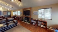 Located at 5001 College St SE in Lacey, Washington, this single-floor space offers a warm and functional layout designed for comfort and practical living. The main living area is expansive and open, featuring rich hardwood-style flooring, a striking stone fireplace with a built-in wall-mounted television above it, and track lighting that complements the ceiling fan overhead. Multiple windows fitted with horizontal blinds allow generous natural light to fill the room, creating a bright and welcoming atmosphere. Leather seating, well-stocked bookshelves, and a cozy reading nook add to the lived-in character of the space.

A secondary seating area with soaring ceilings and warm earth-toned walls provides an inviting lounge space, anchored by a large leather sofa, coordinating accent chairs, and tasteful wall art. This area transitions naturally into the rest of the floor plan, which also includes a dining area with a wooden table and chairs.

The kitchen features granite-style countertops, dark wood cabinetry, tile flooring, and standard appliances including a range, refrigerator, and microwave. A tall pantry cabinet adds additional storage. The overall design of the space blends functionality with a cozy, residential ambiance, making it well-suited for community or shared use.