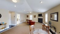 Located at 3150 Boardwalk Ln in Greenville, NC, this single-floor residence offers a welcoming and functional layout designed for comfortable everyday living. The main living area is defined by soaring vaulted ceilings that lend a sense of openness and volume to the space. A ceiling fan is centrally positioned, contributing to both comfort and airflow throughout the room. The living room features a classic fireplace as a focal point, flanked by a wall-mounted television, creating an inviting entertainment hub. Neutral-toned carpeting and warm beige walls give the space a soft, cohesive aesthetic. Natural light filters in through multiple windows fitted with plantation shutters, enhancing the bright and airy atmosphere. A dining area with a round table and wooden chairs is situated nearby, ideal for casual meals. Recessed lighting complements the natural light throughout. An adjacent room, visible through a doorway, appears to serve as a kitchen or breakfast nook, extending the functional footprint of the home. Decorative wall art adds character to the living space. Overall, the home presents a clean, well-maintained interior that balances practicality with a relaxed residential charm.