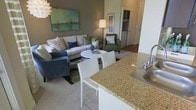 This single-floor space showcases a thoughtfully designed open-concept layout that promotes a sense of connectivity and ease of movement throughout. The kitchen is outfitted with granite countertops featuring warm, speckled tones and a stainless steel sink, providing both functionality and visual appeal. A breakfast bar with seating extends from the counter, creating a casual dining option that bridges the kitchen and living areas.

The living room is warm and inviting, anchored by a light-toned sectional sofa accented with decorative pillows in blue and green hues, complemented by a pair of plush green armchairs. A round white coffee table serves as a central focal point, while a soft area rug defines the seating zone. A floor lamp provides warm ambient lighting, and a large abstract artwork in muted greens and blues adorns the wall, contributing to the space's cohesive color palette.

The neutral, earthy wall tones throughout create a calm and welcoming atmosphere, while the carpeted flooring adds comfort underfoot. A hallway visible in the background hints at additional rooms, including what appears to be a laundry or utility area. The overall ambiance is modern yet approachable, with a clean, coordinated aesthetic suited for comfortable everyday living.