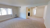 This single-floor space features an open layout with a generously sized living area finished with neutral-toned wall-to-wall carpeting that contributes to a clean and understated aesthetic. The walls are painted in a soft white, creating a bright and airy atmosphere throughout. Natural light enters through a window along one wall, complementing the overhead lighting and enhancing the sense of openness.

A partial dividing wall separates the living area from the kitchen, which is visible through an open passage. The kitchen features warm-toned wood cabinetry and includes a dark-colored range, with light-colored flooring that contrasts with the carpeted living spaces. A floor-level HVAC vent is positioned near the living area's exterior wall, indicating a centralized climate control system.

A ceiling fan with a light fixture is installed in an adjacent carpeted room, visible beyond the kitchen pass-through area. A hallway extends further into the unit, connecting additional rooms and suggesting a functional flow throughout the space. The overall ambiance is neutral, practical, and move-in ready, offering a versatile blank canvas.