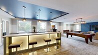 Located at 110 Talisman Way in Raleigh, NC, this thoughtfully designed amenity space offers a sophisticated and versatile environment for socializing and recreation. The layout encompasses a generously sized single floor, seamlessly connecting multiple distinct areas.

The main lounge area commands attention with a bold, geometric blue accent wall housing a large flat-screen TV, complemented by an assortment of plush seating arrangements including barrel chairs and sofas—ideal for gathering and relaxing. An adjoining bar area features an illuminated counter with built-in under-lighting, pendant globe lights suspended from a striking blue ceiling, and bar stools, creating an upscale, social atmosphere.

Natural light pours in through large windows overlooking both the surrounding greenery and the resort-style swimming pool, with granite-topped cabinetry below providing additional storage and counter space. A dedicated game room includes a full-size pool table, while a separate private room—outfitted with wainscoting wall paneling, a wall-mounted TV, and a large poker table—offers a more intimate setting.

A comfortable sitting room with a dark navy ceiling, barn-style sliding glass door, and curated artwork provides a quieter retreat within the space. Throughout, consistent design elements such as board-and-batten wainscoting, recessed lighting, and patterned carpeting unify the space with a modern yet inviting aesthetic.