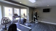 This single-floor fitness room offers a functional and well-equipped space designed to support a variety of workout routines. The room features dark hardwood-style flooring that contrasts cleanly with the neutral grey walls, creating a modern and focused atmosphere. A textured ceiling with recessed lighting and a skylight panel ensures the space is well-lit, while a large window along one wall invites natural light and provides outdoor views, enhancing the overall openness of the room. The cardio section includes a treadmill and an elliptical trainer, positioned to take advantage of the natural light from the window. A multi-functional strength training machine is situated along the opposite wall, offering options for a range of resistance exercises. A wall-mounted flat-screen television is also present, allowing users to stay entertained during workouts. The clean white baseboards and trim provide a polished finish that complements the darker tones of the flooring and walls. Overall, the space is practical, tidy, and thoughtfully arranged to maximize usability within its footprint.