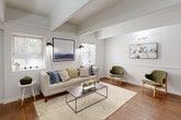 This well-appointed single-floor residence combines classic architectural details with a clean, modern aesthetic. The inviting entryway opens into a bright living area defined by exposed beam ceilings and rich hardwood floors. Large windows on multiple walls flood the space with natural light, creating an airy and welcoming atmosphere. The living room offers generous proportions, with a tufted sofa, mid-century accent chairs, and curated artwork contributing to the space's refined character.

The open layout connects the living area to a dedicated dining nook, complete with a round white table and Windsor-style chairs, and leads directly into the kitchen, which features white cabinetry and full-size appliances. A convenient in-unit laundry closet adds practical functionality.

The primary bedroom is a calm retreat, featuring carpeted floors, exposed beams, and a window with greenery views, lending the room a serene quality. A second flexible room with high vaulted ceilings, carpet, and abundant natural light serves well as an additional bedroom or home office, complete with a built-in shelving unit and multiple seating options.

Throughout the residence, white wainscoting, neutral walls, and consistent design choices create a cohesive and polished living environment that is both comfortable and stylish.