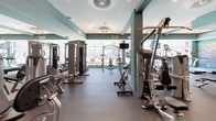 This fitness center is thoughtfully designed to support a comprehensive workout experience. The space features a refined color palette of teal-accented walls paired with crisp white trim and ceilings, creating an energizing yet clean atmosphere. Rubber flooring throughout provides durability and comfort underfoot during exercise.

A substantial collection of Hoist brand strength-training machines lines the perimeter and interior, offering users a wide range of resistance and weight equipment options. Cardio equipment, including stationary bikes, is strategically positioned to take advantage of the floor-to-ceiling windows along the front of the space, allowing natural light to pour in while providing views of the surrounding exterior. Mounted monitors are available near select equipment for entertainment during workouts.

Recessed and flush-mounted ceiling lighting supplements the abundant natural light, ensuring the space remains well-lit throughout the day. Structural columns are integrated into the layout without disrupting the open floor plan. The overall ambiance is modern and professional, providing a clean, organized environment well-suited for a focused fitness routine.