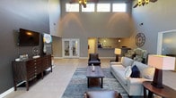 This single-floor communal space is defined by its dramatic double-height ceilings in the main lounge, where a row of clerestory windows floods the interior with natural light and draws the eye upward. Ceiling fans mounted at various heights enhance airflow across the expansive open floor plan, which is finished in large-format tile throughout the primary areas. The main lounge is thoughtfully furnished with a plush sofa, leather accent chairs, and a solid wood coffee table anchored by a patterned area rug, creating a welcoming and comfortable gathering environment. A large flat-screen television and a media console line one wall, while French doors lead to an adjacent office or meeting room. A café-style counter with pendant lighting and a coffee station is visible at the rear, providing a convenient amenity for visitors. Transitioning into the dining area, the space shifts to warm hardwood-style flooring beneath a distinctive paneled wood ceiling with exposed beams and recessed lighting. Large picture windows and glass doors wrap around this room, offering sweeping views of the landscaped grounds, mature trees, and outdoor seating areas beyond. A high-top communal table with industrial-style wood-and-metal stools occupies a windowed nook, ideal for casual meetings or collaborative use. A separate round dining table with upholstered chairs provides additional seating. Together, the areas form a cohesive, light-filled community hub.
