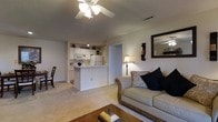 This single-floor residence offers a warm and inviting atmosphere defined by an open-concept layout that seamlessly connects the living area and kitchen. The living room is comfortably appointed, featuring plush carpeting underfoot, a classic ceiling fan with integrated lighting, and soft neutral wall tones that lend a calm, airy feel to the space. An elegant sofa with decorative pillows anchors the seating area, complemented by a wooden coffee table and a classic floor lamp that adds a warm glow. A framed wall mirror and decorative wall sconces add tasteful character to the room.

The kitchen is efficiently designed with white cabinetry, white appliances including a refrigerator and built-in microwave, and a peninsula-style breakfast bar that doubles as a casual dining space and provides a natural dividing point between the kitchen and living areas. Recessed lighting illuminates the kitchen workspace, and charming decorative accents above the cabinets add a personal touch. A doorway visible from the living room leads to additional areas of the residence, suggesting a functional floor plan with private spaces set apart from the main living zone. Overall, the space presents a clean, cohesive aesthetic with a comfortable and practical design.