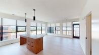 This single-floor unit showcases a thoughtfully designed open-concept layout with a strong emphasis on natural light and elevated views. The living and dining area is expansive, anchored by a wall of black-framed windows that frame panoramic outdoor vistas, allowing daylight to flood the light hardwood floors that run seamlessly throughout the space.

The kitchen is outfitted with floor-to-ceiling white cabinetry, a textured white subway tile backsplash, and stainless steel appliances including a built-in microwave, electric range, dishwasher, and refrigerator. A substantial kitchen island with warm wood cabinetry and a neutral stone countertop serves as the kitchen's centerpiece, illuminated by two sleek cylindrical pendant lights.

The bedrooms are generously sized, each featuring large black-framed windows with roller shades that capture open views of the surrounding cityscape and beyond. The rooms maintain the same light hardwood flooring and clean, neutral palette found throughout.

The bathrooms are finished with hexagonal tile flooring, open wood-framed vanities with integrated sinks, large mirrors, and clean white walls. One bathroom includes a glass-enclosed walk-in shower, while another features a more compact configuration. The overall aesthetic is clean, modern, and cohesive.
