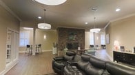 Located at 146 Yester Oaks Dr in Mobile, Alabama, this single-floor community space offers a welcoming and well-appointed environment ideal for gatherings and leisure. The centerpiece of the main room is a dramatic floor-to-ceiling brick fireplace, which anchors the space and adds a classic, rustic charm to the overall aesthetic. Warm earth-toned walls, crown molding, and rich hardwood flooring contribute to a refined yet comfortable atmosphere throughout.

The open layout is thoughtfully arranged to accommodate a variety of uses. A seating area near the fireplace features plush leather reclining sofas and accent chairs, creating a cozy lounge setting. Decorative columns define the space without enclosing it, maintaining an airy, expansive feel. Overhead, large drum pendant lights hang from the ceiling, which features a distinctive circular tray detail that adds architectural interest.

Natural light filters in through multiple windows along the perimeter, brightening the space and offering views of the surrounding exterior. Additional seating areas with bar-height tables and chairs are positioned toward the edges of the room, providing flexible options for casual dining or conversation. The overall ambiance is warm, functional, and inviting, making it well-suited for community events and everyday social interaction.