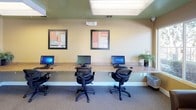 This single-floor business center is designed to provide a functional and comfortable environment for work and collaboration. The space is finished with warm beige walls, a distinctive green-painted ceiling, and neutral carpet flooring throughout, creating a professional yet welcoming atmosphere.

The primary workstation area features a long granite-topped counter running along two walls, accommodating multiple desktop computers with ergonomic mesh chairs. Framed artwork and a wall clock add a polished touch above the workstations. An additional counter section houses a multifunction printer, a desktop computer, and supplemental workspace, ensuring all essential office needs are met in one place.

A dedicated lounge area near the entrance provides comfortable dark leather seating, including armchairs and a sofa arranged around a wooden coffee table. Potted indoor plants add a natural, refreshing element to the space. Large windows along one wall allow natural light to filter in, offering views of the landscaped exterior.

Access to the center is through a glass-paneled door, and the space benefits from recessed and overhead fluorescent lighting for consistent illumination throughout. Informational postings on the walls keep users informed of business center guidelines. Overall, this space offers a complete, self-contained environment suited for computing, printing, and quiet work tasks.