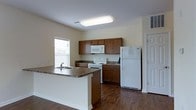 This single-floor residence offers a well-designed, functional layout centered around an open-concept kitchen and living area. The kitchen is a standout feature, showcasing warm medium-toned wood cabinetry that provides ample storage space along both the upper and lower walls. A central kitchen island with a dark laminate countertop and a built-in double sink serves as a practical workspace and natural gathering point, separating the kitchen from the adjacent living area. White appliances, including a refrigerator, range, and over-the-range microwave, complement the clean, neutral aesthetic of the space. The flooring throughout is a rich hardwood-style surface that adds warmth and continuity to the open floor plan. Light-toned walls keep the interior bright and airy, enhanced further by natural light streaming in through a window near the kitchen. Recessed and flush-mounted ceiling lighting ensures the space is well-lit. The overall ambiance is clean, practical, and inviting, making it well-suited for everyday living and casual entertaining. A doorway visible near the refrigerator hints at additional rooms, contributing to the home's functional and accessible single-floor layout.