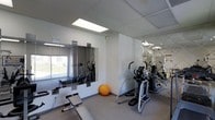 This single-floor fitness space is thoughtfully designed to maximize utility within a streamlined layout. The room features a neutral color palette of white walls and light grey carpeted flooring, creating a clean and focused environment conducive to exercise. A drop ceiling with recessed panel lighting ensures consistent, even illumination throughout the space.

Large mirrored wall panels are a standout feature, positioned along two walls to visually expand the room and allow users to monitor their form during workouts. A window on one wall introduces natural light, adding brightness and an open feel to the interior.

The space is equipped with a variety of cardio machines, including elliptical trainers and what appears to be a treadmill, all positioned to allow comfortable use. A weight bench and additional strength training accessories are visible, offering versatility for different workout routines. An exercise ball adds a functional element for core training.

Overall, the fitness room presents a practical and well-organized setup, suitable for a range of cardiovascular and strength-training activities, with an atmosphere that is both functional and inviting.