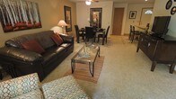 This single-floor residence offers an open-concept floor plan where the living and dining areas flow naturally into one another, creating a cohesive and functional shared space. The living area is anchored by a dark leather sofa accented with rich burgundy throw pillows, paired with a glass-top coffee table resting on a striped area rug. Warm beige carpeting extends throughout, adding a sense of comfort underfoot. A ceiling fan with integrated lighting provides both ventilation and ambient illumination overhead.

Adjacent to the living area, the dining space features a dark wood table surrounded by matching upholstered chairs, set atop a patterned area rug that helps define the zone within the open layout. Framed artwork on the warm-toned walls adds visual interest without overwhelming the space. A floor lamp near the sofa provides additional task lighting.

Beyond the main living and dining area, a glimpse into the background reveals additional seating — possibly a breakfast bar or kitchen nook — suggesting further functional space. The overall ambiance is warm and inviting, characterized by neutral earth tones, coordinated furnishings, and a practical, comfortable layout suited for everyday living.
