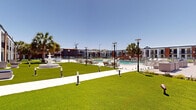 This two-story resort-style property is designed with a contemporary coastal aesthetic, blending bold navy blue and warm wood-tone exterior accents with crisp white facades. The building wraps around a generously sized central courtyard, creating a cohesive outdoor living environment bathed in natural sunlight. At the heart of the grounds lies a large swimming pool enclosed by glass fencing and surrounded by an expansive pool deck furnished with lounge chairs, providing ample space for relaxation and recreation. Manicured green lawns extend throughout the courtyard, punctuated by low-profile pathway lighting and a decorative classical fountain that adds an elegant focal point to the landscape. Tall palm trees are strategically placed throughout the grounds, enhancing the tropical ambiance. Ground-floor units open directly onto private outdoor seating areas furnished with contemporary rattan lounge sets, while second-floor units feature individual balconies with metal railings, offering elevated views of the lush courtyard. A covered pavilion structure with a distinctive blue metal roof serves as an additional communal gathering space. The overall atmosphere is bright, open, and resort-like, combining modern architectural design with inviting outdoor amenities suited for an active and leisurely lifestyle.