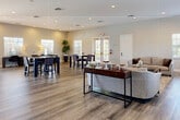 The Greens Edge Lobby Clubhouse in Fort Myers, Florida is a thoughtfully designed single-floor community space that offers a warm and inviting atmosphere for residents and visitors alike. The expansive open floor plan is anchored by wide-plank wood-look flooring that flows seamlessly throughout, complemented by neutral walls and recessed lighting that create a bright, clean aesthetic.

Upon entry, guests are welcomed by a striking feature wall covered in lush greenery, accented by a vibrant pink neon "Welcome Home – Greens Edge" sign, setting a distinctive and memorable tone. Adjacent to the entry is a professional leasing office enclosed in sleek glass partitions, complete with a seating area and a detailed community map on display.

The main gathering area is generously proportioned, accommodating multiple functional zones. A comfortable lounge area features a neutral sofa, armchairs, and a coffee table arranged on a plush area rug, alongside a mounted flat-screen television and natural light streaming through plantation-shuttered windows. Nearby, bar-height dining tables with upholstered chairs provide space for casual dining or socializing.

A dedicated activity and workspace zone offers additional high-top tables and a computer workstation. The space benefits from abundant natural light through multiple French doors and large windows, with views of the surrounding outdoor greenery. The overall design balances functionality and comfort, making it an ideal hub for community life.