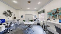 Located at 5009 Park Central Dr in Orlando, FL, this single-floor community business center offers a thoughtfully designed space for work and collaboration. The layout flows seamlessly from an elegant reception lounge into a functional business suite. The lounge area sets a refined tone with a tray ceiling, a statement chandelier, large floor planters with tropical greenery, plush white armchairs, a colorful area rug, and glass-paneled French doors that provide a visual connection to the work area beyond. The business suite is finished with slate-tiled flooring in a classic checkerboard pattern and is lined with white walls accented by crown molding. Multiple computer workstations with adjustable bar-height seating are arranged along the perimeter, complemented by a multifunction printer and copier for full office capability. A round white table with modern wire chairs serves as a central gathering point, suitable for informal meetings or group work. Large windows dressed with patterned valances allow natural light to fill the room while offering views of the surrounding landscape. Decorative wall art and potted plants add warmth to the otherwise crisp, professional aesthetic. Recessed lighting throughout ensures consistent illumination across the entire space.