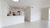 This single-floor unit showcases a contemporary open-concept design defined by a crisp, all-white color palette and light wood-style flooring that runs seamlessly throughout the living and kitchen areas. The space is bright and airy, with flush-mount and track lighting providing consistent illumination across the interior.

The kitchen is integrated into the main living area via a white peninsula counter, offering both workspace and a natural divider between the cooking and living zones. Stainless steel appliances, including a french-door refrigerator with an ice and water dispenser, are visible within the kitchen, complemented by white cabinetry above. A stacked washer and dryer unit is also visible through a pass-through or hallway, indicating in-unit laundry. Track lighting over the kitchen adds a modern, functional touch.

The open living area features clean white walls, crisp white baseboards, and a central HVAC vent, highlighting the unit's modern construction. The uncluttered, minimalist design creates a sense of openness, allowing for flexible furniture arrangements. A hallway visible in the background suggests additional rooms beyond the main living space.