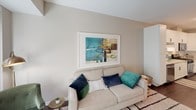 This well-appointed single-floor residence offers a thoughtfully designed layout that balances comfort and modern style. The living area is bright and welcoming, anchored by a large sliding glass door that opens onto a private balcony, flooding the space with natural light and offering lush, tree-lined views. The balcony provides an intimate outdoor retreat, complete with space for seating and al fresco dining.

The open-concept kitchen is a standout feature, boasting crisp white shaker cabinetry, granite countertops, a subway tile backsplash, and a full suite of stainless steel appliances including a gas range, microwave, dishwasher, and refrigerator. A central kitchen island with bar seating adds both functionality and a casual dining option. Rich dark hardwood flooring flows throughout the kitchen and main living areas, lending warmth and continuity.

A stacked washer and dryer unit is conveniently integrated within the residence, adding everyday practicality. The bathroom features a granite-topped vanity, a clean walk-in shower, and modern fixtures that echo the home's contemporary aesthetic.

The bedroom is a tranquil retreat, carpeted for comfort and fitted with large windows that invite natural light and treetop views. A ceiling fan adds to the relaxed ambiance of the room. Neutral tones throughout the residence create a cohesive, serene atmosphere.
