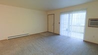 This single-floor unit features a spacious living area finished with wall-to-wall carpeting in a neutral beige tone, lending a clean and understated aesthetic throughout the space. The room is bathed in natural light from a large sliding glass door fitted with vertical blinds, which opens to an outdoor area — providing both brightness and convenient exterior access. A front entry door with a brown trim frame offers a clear and functional entryway into the space. Along the base of the wall, a baseboard heating unit ensures efficient climate control during cooler months. The walls are finished in a soft, off-white tone, creating a bright and open atmosphere that is well-suited for a variety of furnishing arrangements. The straightforward, practical layout of this single-level space makes it easy to configure for everyday living. The combination of natural light from the sliding door, neutral finishes, and a generous open floor plan gives the space a comfortable and welcoming character, ideal for those seeking a simple and functional living environment.