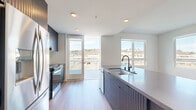 This modern San Diego residence offers a sleek, contemporary design across a single floor, accommodating five bedrooms and three bathrooms. The open-concept kitchen and living area serves as the heart of the home, anchored by a striking dark-finished island with an integrated sink and matte countertops. Fluted dark cabinetry contrasts elegantly with white walls and light wood-tone flooring found throughout. Stainless steel appliances, including a full-size refrigerator and range, complete the kitchen. Floor-to-ceiling windows and a glass-railed balcony flood the living area with natural light while framing panoramic views of the surrounding hillside landscape. Each bedroom features large windows that continue the theme of natural light and outdoor connectivity, with several rooms offering generous closet space, including walk-in configurations with dual hanging rails. The three bathrooms are finished in clean white tiling with dark wood vanities, maintaining a consistent modern aesthetic. A central hallway connects all rooms efficiently. The overall ambiance is bright, minimalist, and contemporary, reflecting a thoughtfully curated design suited to modern San Diego living.