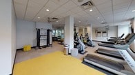This fitness center is a comprehensive, single-floor facility designed to support a full range of workout routines. The space is organized into distinct functional zones, making it easy to navigate between cardio, strength training, and flexibility exercises.

The cardio section is lined with Matrix treadmills and elliptical machines positioned along a wall of large, black-framed windows that allow ample natural light to fill the room and provide outdoor views. Wall-mounted flat-screen televisions accompany the cardio equipment for entertainment during workouts.

The strength training area features a multi-station cable machine along with free weights, benches, and additional resistance equipment, all reflected in large wall mirrors that enhance the sense of openness. Instructional workout charts are posted on the wall for user reference.

A dedicated stretching and floor exercise zone is defined by vibrant yellow flooring, offering a visually distinct area for yoga mats, stability balls, and bodyweight training. Ceiling fans and recessed lighting provide comfortable climate control and even illumination throughout.

The facility also includes water stations conveniently located near the entrance corridor, along with waste receptacles throughout. The drop ceiling with recessed lighting and neutral wall tones give the space a clean, modern aesthetic suited to a professional fitness environment.