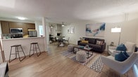 This single-floor residence offers a thoughtfully designed open-concept layout that seamlessly connects the living, dining, and kitchen areas. The living room is a welcoming space anchored by a charcoal sofa, a geometric-patterned area rug, and a large abstract canvas that adds a vibrant pop of color to the neutral walls. Light wood-look flooring runs throughout, complementing the bright, airy atmosphere. A large sliding door with vertical blinds allows natural light to filter in and provides access to the outdoors.

The dining area sits adjacent to the living space and features a round white table with woven chairs beneath a ceiling fan, creating a casual yet stylish setting. A mirrored closet behind the dining area adds depth and reflects light throughout the room.

The kitchen is equipped with stainless steel appliances, dark wood cabinetry, and a generous white countertop that doubles as a breakfast bar, ideal for quick meals or entertaining.

The bedrooms are comfortably sized, each featuring light wood flooring, natural window views of surrounding greenery, mirrored sliding closet doors, and clean neutral walls that create a calm, restful environment.

The bathroom is crisp and functional, featuring a wide vanity with a large mirror, light countertops, and a shower-tub combination. Textured ceilings are consistent throughout the home, adding a classic residential character to the overall space.