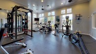 This fitness center is a thoughtfully designed, single-level workout facility that offers a comprehensive range of equipment for both cardio and strength training. The space is bright and inviting, with large windows lining the walls that flood the room with natural light and provide views of the surrounding outdoor area, including what appears to be a pool and landscaped grounds.

The flooring throughout is durable rubber, ideal for a high-use fitness environment. Recessed lighting and a ceiling fan complement the natural light, ensuring the space remains comfortable and well-lit during any time of day. Crown molding adds a refined architectural touch to the otherwise functional interior.

The cardio section is well-stocked with treadmills, Cybex stationary bikes, elliptical trainers, and a air resistance bike, all positioned to take advantage of the outdoor views through the large windows. A wall-mounted television is also present for entertainment during workouts.

The strength training area features a full power rack with a barbell and weight plates, a cable machine system, a dumbbell rack with a full set of free weights, an adjustable bench, and a plyo box. Full-length mirrors line one wall, enhancing the sense of space and allowing users to monitor their form.

Additional equipment such as a battle rope, a speed bag, and a water cooler round out the facility's offerings. A separate doorway leads to adjacent rooms, suggesting access to restrooms or other amenities nearby.