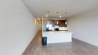 This well-appointed 1-bedroom, 1-bathroom unit is designed with a clean, modern aesthetic that emphasizes open living and functional design. The open-concept layout seamlessly connects the living area to the kitchen, creating a spacious and airy feel throughout the main living space. Durable hardwood-style flooring flows continuously across the entire unit, adding warmth and cohesion to the contemporary interior.

The kitchen serves as a central highlight, featuring a large island with a crisp white countertop that provides ample prep space and doubles as a casual dining area. Rich wood-toned cabinetry with a flat-front design lines the upper and lower walls, complemented by stainless steel appliances including a French door refrigerator, microwave, and range. Pendant lighting hangs elegantly above the island, while recessed lighting throughout ensures the space is well-lit.

Neutral white walls and smooth ceilings give the unit a bright, clean atmosphere, while the open hallway leading to the bedroom and bathroom offers a sense of depth. The thoughtful single-floor layout maximizes ease of living, with each area flowing naturally into the next. Overall, this unit delivers a polished, contemporary living experience with quality finishes at every turn.