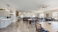 This well-appointed community clubhouse offers a generous, open-concept layout spread across a single floor. The interior is finished with durable wood-look tile flooring throughout, crown molding, and recessed lighting, all contributing to a clean and welcoming ambiance. Walls feature a distinctive two-tone design with soft blue wainscoting beneath neutral upper walls, adding a cohesive and polished aesthetic.

The lounge area serves as a central gathering point, anchored by a large curved sectional sofa and a striking brick fireplace with a wall-mounted television and rustic open shelving. Ceiling fans are positioned throughout the space to ensure comfort. Adjacent to the lounge, expansive dining areas accommodate multiple large tables and chairs, ideal for group gatherings and community events. Abundant windows allow natural light to flood the space and offer outdoor views of the surrounding green landscape.

The fully equipped kitchen features gray shaker-style cabinetry, white quartz countertops with a waterfall island, subway tile backsplash, stainless steel appliances, and pendant lighting — designed for functional communal use. A window above the counter provides a direct view of an outdoor playground and pool area. French doors and multiple entry points provide convenient access to the outdoors, completing this versatile and inviting community space.