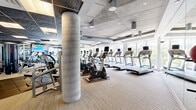Located at 500 West Ave in Norwalk, Connecticut, this fitness center offers a comprehensive and well-appointed exercise space spread across a single floor. The facility is designed to accommodate a full range of workout routines, with distinct zones dedicated to cardiovascular training and strength work.

The cardio area features a row of treadmills, elliptical machines, and stationary bikes, all equipped with individual display screens. Large floor-to-ceiling windows line the perimeter, flooding the space with natural light and providing views of the surrounding neighborhood. Full-length mirrors along the walls visually expand the room and assist users during exercise.

The strength training zone includes a robust selection of free weights, a complete dumbbell rack, cable and multi-functional resistance machines, and rubber-floored lifting platforms that provide both safety and durability. An overhead decorative tiled ceiling with recessed lighting adds a refined aesthetic contrast to the exposed industrial-style ductwork visible in other sections of the space.

The flooring transitions between carpet-textured material in the cardio zone and hard wood-toned surfaces in the strength area, clearly delineating functional spaces. The overall ambiance is modern and professional, combining industrial architectural details with upscale finishes to create an inviting and fully functional fitness environment.
