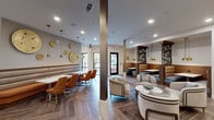 Located at 2900 N Braeswood Blvd in Houston, Texas, this single-floor community lounge is a thoughtfully designed social and gathering space that blends contemporary style with functional versatility. The open layout is unified by striking herringbone-patterned wood-look tile flooring that flows seamlessly throughout, anchoring the sophisticated interior design.

The lounge is organized into several distinct zones. A casual seating area features plush curved armchairs arranged around a marble-top round table, ideal for relaxed conversation. Adjacent to this is a dining area with warm caramel-toned banquette seating paired with cognac leather chairs and white tables, creating an inviting café-like atmosphere. Decorative laser-cut wood panels serve as stylish dividers between the booth seating sections, adding architectural interest and privacy.

A built-in coffee and refreshment bar is equipped with countertop appliances, a sink, and blue-grey cabinetry with marble countertops, complemented by open shelving displaying curated décor. A dedicated work area with bar-height seating and pendant lighting provides a focused workspace option.

Natural light fills the space through large black-framed glass doors and windows that open directly to an outdoor area, offering views of the surrounding grounds. Gold pendant lights, metallic wall art, and warm recessed lighting complete the polished, upscale ambiance throughout.