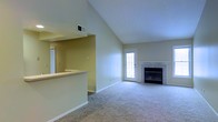 This single-floor residence offers an inviting and well-designed layout that balances openness with functional living. The main living area immediately draws attention with its dramatic vaulted ceilings, which lend a spacious, airy quality to the room. Soft, neutral-toned carpeting extends throughout the living space, contributing to a warm and comfortable ambiance. A tile-surround fireplace serves as a natural focal point, providing both aesthetic charm and a cozy gathering spot. Natural light filters into the room through a classic multi-pane exterior door, brightening the interior and creating a welcoming atmosphere. Adjacent to the living area, a pass-through counter connects to the kitchen, promoting an open and social flow between spaces. The kitchen features a raised breakfast bar-style counter, ideal for casual dining or entertaining. Neutral wall tones throughout the space complement the architectural elements and provide a versatile backdrop for a variety of interior styles. The ceiling includes a centrally positioned HVAC vent, reflecting thoughtful climate control integration. Overall, this residence combines practical design with understated elegance, making it a comfortable and functional single-floor living space.