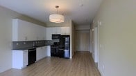 This single-floor residential unit offers a clean, modern aesthetic anchored by an open-concept layout that seamlessly connects the kitchen and living areas. The kitchen is well-appointed with crisp white upper and lower cabinetry, dark countertops, and a full suite of black appliances including a gas range, microwave, and refrigerator. A stylish drum pendant light hangs above, providing warm, focused illumination. A tile backsplash adds a subtle textural detail behind the range, while the under-mount sink is positioned along the window-side counter, lending a practical and polished look. Light hardwood-style flooring runs continuously throughout the space, contributing to a bright and cohesive feel. The open corridor leads directly to a modern entryway door, finished in a warm wood tone that provides a welcoming contrast to the neutral wall palette. The walls are painted in a soft, muted tone that complements the light flooring and white trim. The overall ambiance is contemporary and functional, with a layout designed to maximize usability and flow. The space presents a well-balanced combination of form and function, making it an inviting and practical living environment.