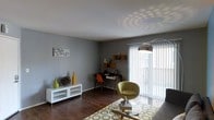 This single-floor space showcases a thoughtfully designed living area that blends modern and eclectic styles. The room is anchored by rich, dark hardwood flooring that contrasts elegantly with the neutral grey walls, creating a contemporary and cohesive aesthetic throughout.

The living area is well-lit, benefiting from a large sliding glass door dressed with vertical blinds that allows abundant natural light to filter in while offering privacy control. A stylish arc floor lamp with a metallic shade adds a modern touch and contributes to the ambient lighting, casting distinctive circular patterns across the white ceiling.

The space is versatile, accommodating both a lounging area and a dedicated workspace. The sitting area features a distinctive olive-green accent chair paired with a glass-top coffee table and a patterned area rug in green and white tones. A sleek white media console serves as a functional yet decorative storage piece along one wall.

A compact desk setup in the corner, complete with an orange mid-century modern chair and open shelving displaying decorative objects, adds a functional dimension to the room. The overall ambiance is casual yet curated, with carefully selected décor accents in warm yellows and greens providing pops of color against the cool grey backdrop.