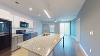 This modern single-floor residence showcases a thoughtfully designed open-concept layout that seamlessly connects the kitchen and living areas. The kitchen is a standout feature, equipped with a large granite-topped island with an integrated sink, two-tone cabinetry combining white upper cabinets with gray lower drawers, and a full suite of black appliances including a refrigerator, range, microwave, and dishwasher. Light hardwood flooring flows throughout the main living areas, enhancing the bright and airy atmosphere created by large windows and a sliding glass door that fills the space with natural light. A ceiling fan serves the open living area, which offers direct access to an outdoor balcony. The bedrooms are carpeted for comfort, each featuring windows with roller shades that frame views of surrounding greenery. The bathrooms are finished with large-format marble-style tile on both the floors and tub surround, complemented by clean white fixtures and granite vanity countertops. Accent walls in muted tones of teal and gray add subtle visual interest throughout the unit. The overall design is clean, contemporary, and move-in ready, balancing functional living spaces with modern aesthetic details.