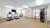 Located at 4727 Arbor Crossing SE in Alexandria, Minnesota, this fitness facility offers a single-floor exercise room designed to support a well-rounded workout routine. The space is finished with durable, neutral-toned carpet flooring and bright white walls, creating a clean and functional environment. Overhead fluorescent lighting ensures the room is well-lit throughout.

The cardio area is equipped with two treadmills and an elliptical machine, positioned near windows that allow natural light to filter in and provide outdoor views. A wall-mounted flat-screen television accompanies the cardio equipment, offering entertainment during workouts. A multi-functional cable resistance machine with yellow accents anchors the strength training zone, providing versatility for a range of exercises. Reference charts are posted on the wall nearby for exercise guidance.

A wall-mounted air conditioning unit helps regulate the room's temperature year-round, complemented by baseboard heating along the perimeter. A built-in dual-level drinking fountain is conveniently mounted on the wall, and a dedicated open area with curtained partitioning offers space for stretching or floor exercises. A wood-trimmed entry door with posted notices provides access to the room, completing this practical and accessible fitness space.