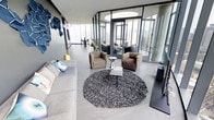 This single-floor high-rise residence exemplifies modern urban living through its striking architectural design and refined interiors. The open-plan layout is anchored by expansive floor-to-ceiling glass walls that wrap around the perimeter, flooding the space with abundant natural light and offering panoramic city skyline views from an elevated vantage point.

The main living area is thoughtfully arranged with a generous sectional sofa, a pair of plush lounge chairs, and a circular patterned area rug that grounds the seating arrangement around a sleek minimalist coffee table. The polished concrete-toned flooring runs seamlessly throughout, reinforcing the space's contemporary character.

A distinctive glass-enclosed room with a circular skylight serves as a dedicated lounge or reading nook, providing an intimate retreat within the larger open space. A dining area with a round table and accent chair is visible beyond, bathed in natural light from the surrounding glazing.

Artistic wall accents, including a bold blue sculptural piece, add visual interest and personality to the otherwise neutral and refined palette. The overall ambiance is one of calm sophistication — airy, luminous, and meticulously curated for both comfort and style, making it an exemplary showcase of elevated contemporary design.