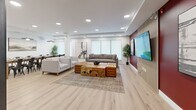 Located at 2165 Benvoulin Crt in Kelowna, BC, this thoughtfully designed amenity lounge offers a welcoming and versatile space for residents to gather, relax, and entertain. The single-level layout flows seamlessly between two distinct zones, unified by light wood-tone flooring and a bright, neutral palette throughout.

The lounge area serves as the heart of the space, anchored by a plush grey sofa and armchairs arranged around a striking live-edge wood coffee table paired with tufted leather ottomans. A bold deep-red feature wall with vertical wood slat accents frames a large wall-mounted television, creating a cozy yet dramatic focal point. Recessed pot lighting illuminates the space evenly, while large windows dressed with roller shades allow in generous natural light.

Adjacent to the lounge, a generously sized dining area accommodates a long wooden communal table surrounded by industrial-style metal chairs, lit by two pendant fixtures that add warmth and character. Curated artwork adorns the walls throughout, enhancing the refined atmosphere.

A sleek kitchenette supports the entertaining function of the space, featuring dark cabinetry with reeded glass upper doors, white quartz countertops, a white subway tile backsplash, stainless steel sink, and a dishwasher — all finished with open shelving for stylish display storage.