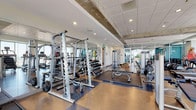 This fitness center on Blount Street in Raleigh, NC offers a thoroughly equipped, single-floor facility designed to support a wide range of workout routines. The space is defined by its modern aesthetic, featuring exposed concrete ceiling beams that contrast with a clean white drop ceiling, recessed lighting, and durable mixed flooring that transitions between hardwood-style and rubberized mat surfaces.

The main workout area is thoughtfully organized into distinct zones. The strength training section houses a comprehensive selection of Life Fitness cable machines, multi-station weight racks, barbells, a full dumbbell rack ranging across multiple weights, adjustable benches, and squat racks. Floor-to-ceiling mirrors line the walls, enhancing the sense of space and allowing users to monitor their form.

The cardio zone is positioned along the perimeter, benefiting directly from floor-to-ceiling glass walls and large sliding doors that open to an outdoor terrace. Treadmills, StairMasters, stationary bikes, and ellipticals are arranged to take full advantage of the panoramic views of the Raleigh skyline.

A dedicated functional training area includes kettlebells, stability balls, a rowing machine, and a Pilates-style reformer, providing versatile options for low-impact and flexibility training. The facility also includes a separate room for additional equipment and private use, along with clearly posted fitness center rules and emergency information near the entrance.