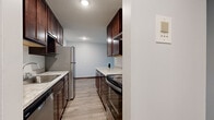 This single-floor unit offers a practical and thoughtfully designed layout, encompassing a combined kitchen and living area along with a full bathroom. The galley-style kitchen is a standout feature, boasting rich dark-wood cabinetry with brushed hardware, light granite countertops, and a full suite of stainless steel appliances including a refrigerator, electric range, and dishwasher. A textured ceiling and recessed lighting add dimension overhead, while light wood-style flooring runs throughout, creating a cohesive and clean aesthetic.

The living area is open and versatile, bathed in natural light from a large window dressed with vertical blinds. The neutral gray walls provide a calm, move-in-ready backdrop, and a baseboard heating unit ensures year-round comfort. The space flows easily from the kitchen, making it well-suited for both everyday living and entertaining.

The bathroom continues the consistent design language of the unit, featuring a dark wood vanity with a white countertop and integrated sink, a large frameless mirror, and matching cabinetry above for added storage. The overall ambiance of this space is clean, modern, and functional, with cohesive finishes tying each area together seamlessly.