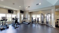 This fitness center offers a clean, functional workout environment designed to accommodate a variety of exercise routines. The space features soaring vaulted ceilings that lend an open, airy feel, complemented by recessed lighting and large windows on multiple walls that flood the room with natural light and provide views of the surrounding greenery outside.

The workout floor is finished with durable dark rubber flooring, ideal for fitness use and easy maintenance. The cardio section is well-stocked, featuring a treadmill, an elliptical machine, and a stationary bike, providing options for a range of aerobic workouts. For strength training, a multi-station cable machine and a leg press/extension unit offer targeted resistance exercises suited to various fitness levels.

A wall-mounted flat-screen television is positioned prominently to keep members entertained during their workouts. The neutral beige walls and white trim give the space a bright, welcoming atmosphere, while the thoughtful layout ensures ample room to move freely between equipment. Overall, this fitness center combines practical amenities with a comfortable, well-lit environment to support an effective and enjoyable exercise experience.