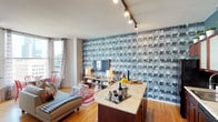 This thoughtfully designed urban residence showcases a bold, eclectic aesthetic across its single floor. The open-concept living and kitchen area serves as the heart of the space, anchored by a striking floor-to-ceiling photographic wallpaper that adds dramatic visual depth. The kitchen features rich wood cabinetry, a mosaic tile backsplash, black appliances, and a generous granite-topped island — ideal for both cooking and entertaining. A teal accent wall and row of potted plants along the upper cabinets lend a fresh, curated feel.

The living area flows naturally toward a curved bay window that frames sweeping city skyline views. Warm hardwood floors, track lighting, and a compact dining nook with vibrant red chairs complete this inviting gathering space.

The bedroom is a standout, featuring a rich crimson damask accent wall adorned with chandelier motifs, offering a glamorous, intimate atmosphere. Large windows frame iconic urban architecture, flooding the room with natural light. An open closet provides convenient storage.

The bathroom is finished in a bold dark palette with striped ceiling detailing, a white vanity, tub-shower combination, and built-in shelving — balancing dramatic style with everyday functionality. Throughout, the residence demonstrates a confident design sensibility with carefully layered textures and colors.