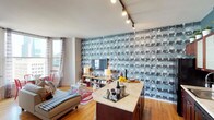 This thoughtfully designed urban residence showcases a bold, eclectic aesthetic across its single floor. The open-concept living and kitchen area serves as the heart of the space, anchored by a striking floor-to-ceiling photographic wallpaper that adds dramatic visual depth. The kitchen features rich wood cabinetry, a mosaic tile backsplash, black appliances, and a generous granite-topped island — ideal for both cooking and entertaining. A teal accent wall and row of potted plants along the upper cabinets lend a fresh, curated feel.

The living area flows naturally toward a curved bay window that frames sweeping city skyline views. Warm hardwood floors, track lighting, and a compact dining nook with vibrant red chairs complete this inviting gathering space.

The bedroom is a standout, featuring a rich crimson damask accent wall adorned with chandelier motifs, offering a glamorous, intimate atmosphere. Large windows frame iconic urban architecture, flooding the room with natural light. An open closet provides convenient storage.

The bathroom is finished in a bold dark palette with striped ceiling detailing, a white vanity, tub-shower combination, and built-in shelving — balancing dramatic style with everyday functionality. Throughout, the residence demonstrates a confident design sensibility with carefully layered textures and colors.