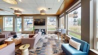 Located at 2448 Winnetka Ave N in Golden Valley, Minnesota, this single-floor community clubhouse offers a well-appointed gathering space designed for both relaxation and socializing. The layout encompasses a welcoming reception area with warm tile flooring, a granite-topped front desk, framed artwork along the walls, and comfortable seating that sets a polished first impression upon entry.

The main lounge area is expansive and thoughtfully arranged with multiple modern seating groupings, including plush sofas and accent chairs positioned around a fireplace and a large wall-mounted television. Distinctive patterned carpeting in rich tones of blue, gold, and brown anchors the space, while an array of drum pendant light fixtures and a coffered ceiling detail with warm wood accents add architectural character. Generous windows line the perimeter, flooding the interior with natural light and offering views of the surrounding landscape and outdoor amenity areas.

A fully equipped kitchen features a granite-topped island with bar seating, stainless steel appliances, warm wood cabinetry, and pendant lighting, making it ideal for entertaining. Adjacent to the lounge, a dedicated game room houses a full-size pool table beneath an industrial-style pendant fixture, with high-top tables positioned near windows overlooking an outdoor pool and patio area. Throughout, the design blends contemporary furnishings with inviting, warm-toned finishes to create a cohesive and functional community space.