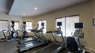 This single-story fitness center is designed to support a well-rounded cardio workout routine. The space is outfitted with multiple Matrix-brand treadmills arranged in a row, alongside an elliptical trainer, providing users with a variety of cardio equipment options. Each treadmill station is paired with a dedicated wall-mounted flat-screen television, allowing users to stay entertained during their workouts. An additional stationary bike is visible toward the rear of the space, further expanding the equipment offerings.

The room features neutral-toned walls and a textured ceiling with recessed lighting that keeps the space well-lit. Large windows fitted with horizontal blinds line the walls, allowing a generous amount of natural light to fill the room and offering views of the surrounding greenery outside. The flooring beneath the equipment appears to be a durable, low-pile carpet suited for a fitness environment.

Overall, the fitness center maintains a clean, functional layout with adequate spacing between machines, creating a comfortable environment for exercise. The combination of natural light, entertainment options, and varied cardio equipment makes this a practical and user-friendly workout facility.