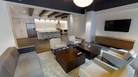 This single-floor space showcases a thoughtfully designed open-concept layout that seamlessly blends a comfortable lounge area with a fully equipped kitchen. The living area features a clean, contemporary aesthetic, with neutral-toned upholstered seating arranged around rich, dark wood coffee tables atop a textured area rug, creating an inviting and functional gathering space. A wall-mounted flat-screen television anchors one end of the room, while a wooden credenza provides additional storage and display space.

The kitchen is a standout feature, boasting a generous island with sleek bar seating, professional-grade stainless steel appliances including a range with a wall-mounted hood, and a panel-ready refrigerator. Warm pendant lighting illuminates the workspace, complemented by subtle under-cabinet lighting that highlights the textured backsplash.

Architectural interest is added through the use of dark exposed wood beams that contrast beautifully against the light ceiling and walls. Wide-plank hardwood flooring flows throughout, unifying the space. The interplay of dark and light tones — from the charcoal ceiling detail to the crisp white walls — gives the space a bold yet sophisticated ambiance well-suited for both everyday use and entertaining.