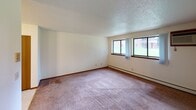 This single-floor residential unit offers a well-organized layout with a clear separation between living, sleeping, and functional spaces. The living room is generously sized, featuring plush wall-to-wall carpeting, baseboard heating, and a pair of windows that invite natural light and outdoor views. A textured ceiling runs throughout the unit, adding a consistent visual element across all rooms. The transition between the living area and kitchen is defined by a shift to light tile flooring. The kitchen is equipped with rich, warm-toned wood cabinetry — both upper and lower — offering ample storage, alongside a double-basin stainless steel sink, a white electric range, and a full-size refrigerator. A ceiling fan with a light fixture provides both ventilation and illumination. The unit includes two bedrooms, each carpeted and featuring sliding-door closets, individual windows with views of surrounding greenery and the exterior, and baseboard heating. The full bathroom includes a combined tub and shower, a vanity with countertop, and tile flooring. Throughout the unit, wood-toned doors and trim provide a cohesive, classic aesthetic, while the neutral wall palette creates a versatile, move-in-ready environment.