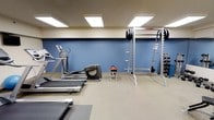This fitness center offers a practical and well-organized space for both cardio and strength training workouts. The room features a neutral color palette with blue accent walls and a light-toned ceiling fitted with recessed panel lighting, ensuring consistent, bright illumination throughout the space. The flooring consists of a durable, textured tile surface well-suited for exercise use.

On the cardio side, the facility includes a treadmill and at least one elliptical trainer, providing options for low-impact and traditional aerobic exercise. For strength training, the space is equipped with a power rack or squat rack loaded with weight plates, along with a dumbbell rack offering a range of free weights. A small step platform is also present, adding versatility for functional fitness routines.

The layout is efficient, allowing users to move between equipment with ease. The clean, uncluttered design keeps the focus on functionality, making this fitness center a convenient and comprehensive option for maintaining an active lifestyle.