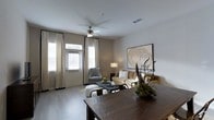 Located at 2170 Benton Blvd in Savannah, Georgia, this single-floor residence offers a thoughtfully designed living space with a cohesive and refined aesthetic. The open-concept layout seamlessly connects the dining and living areas, creating a fluid, functional flow throughout the home. Rich, dark hardwood-style flooring anchors the space, providing a striking contrast to the soft, neutral-toned walls that lend warmth and sophistication to the interior. The dining area features a substantial dark wood table, setting the stage for comfortable gatherings. The adjoining living area is bathed in natural light, courtesy of multiple large windows dressed in elegant floor-to-ceiling cream drapery with dark trim accents. A ceiling fan overhead enhances comfort while contributing to the room's clean, modern character. The living area is further accented by a large decorative wall piece, adding an artistic focal point to the space. A welcoming front entry with a glass-panel door allows additional natural light to filter in. The overall ambiance strikes a balance between understated elegance and everyday livability, making this Savannah residence a well-appointed and inviting home.
